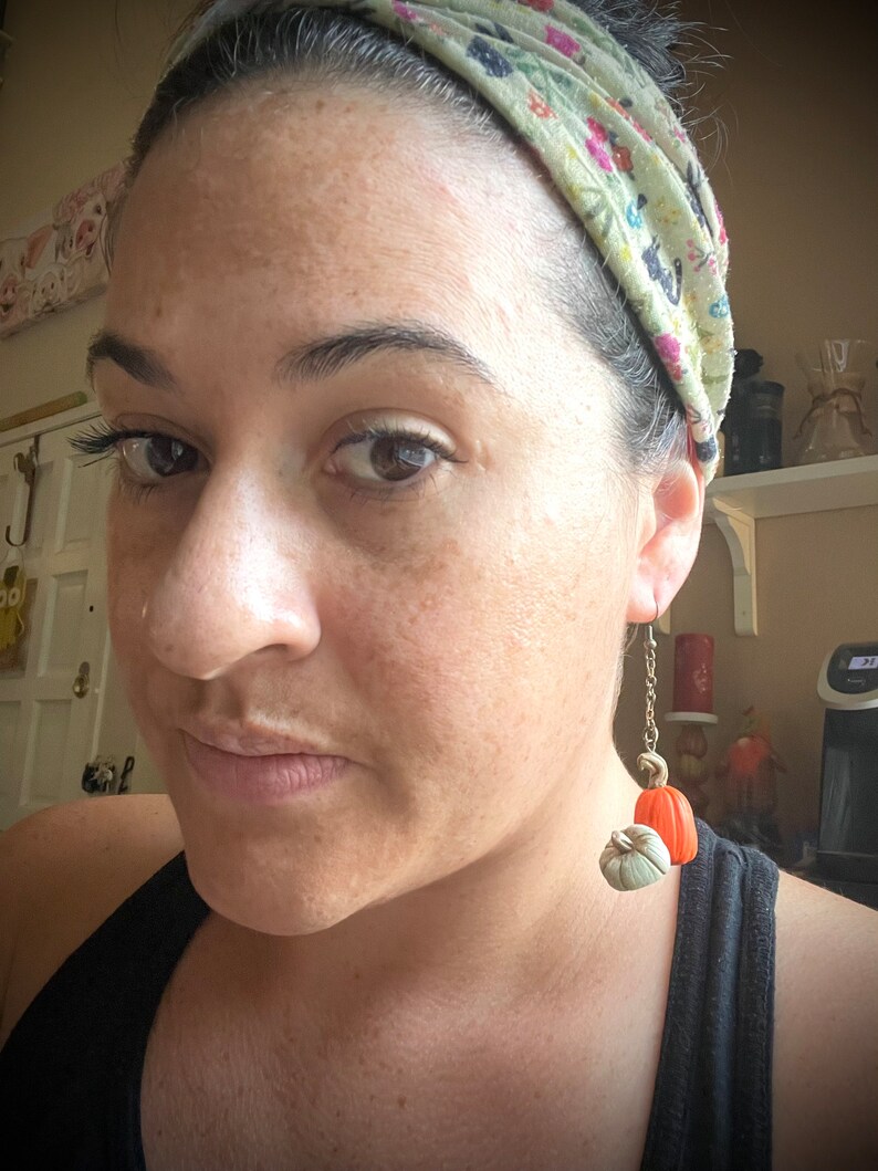 May include: A close-up shot of a person wearing pumpkin-shaped earrings. The earrings feature a small, light green pumpkin and a larger orange pumpkin, suspended from a gold-colored chain. The person is wearing a black tank top and a floral headband.