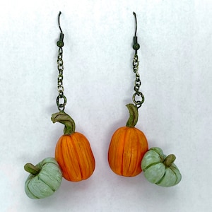 May include: A pair of dangle earrings featuring miniature orange pumpkins with green stems and small, light green pumpkins. The earrings hang from dark green chains and hooks, perfect for fall or Halloween.