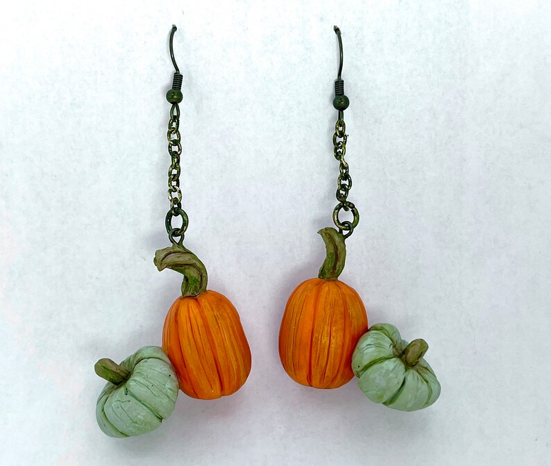 May include: A pair of dangle earrings featuring miniature orange pumpkins with green stems and small, light green pumpkins. The earrings hang from dark green chains and hooks, perfect for fall or Halloween.