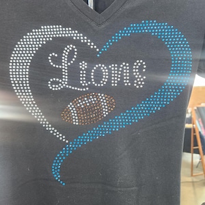 Może przedstawiać: Czarny t-shirt z dekoltem w serek z motywem w kształcie serca. Serce obrysowane jest białymi i niebieskimi kryształkami, a słowo "Lions" zapisane jest białymi kryształkami. Grafika piłki nożnej znajduje się w środku serca.
