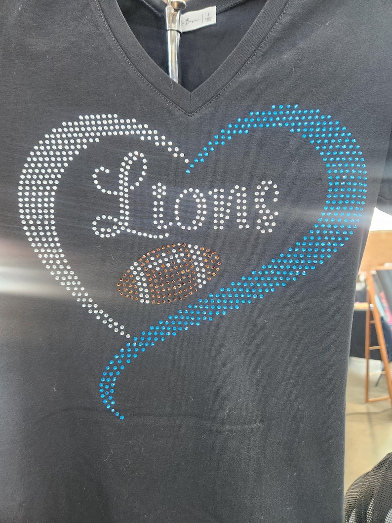 Może przedstawiać: Czarny t-shirt z dekoltem w serek z motywem w kształcie serca. Serce obrysowane jest białymi i niebieskimi kryształkami, a słowo "Lions" zapisane jest białymi kryształkami. Grafika piłki nożnej znajduje się w środku serca.