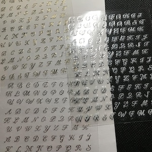 May include: Three sheets of clear plastic alphabet stickers with a cursive font. The first sheet has gold letters, the second sheet has silver letters, and the third sheet has black letters.