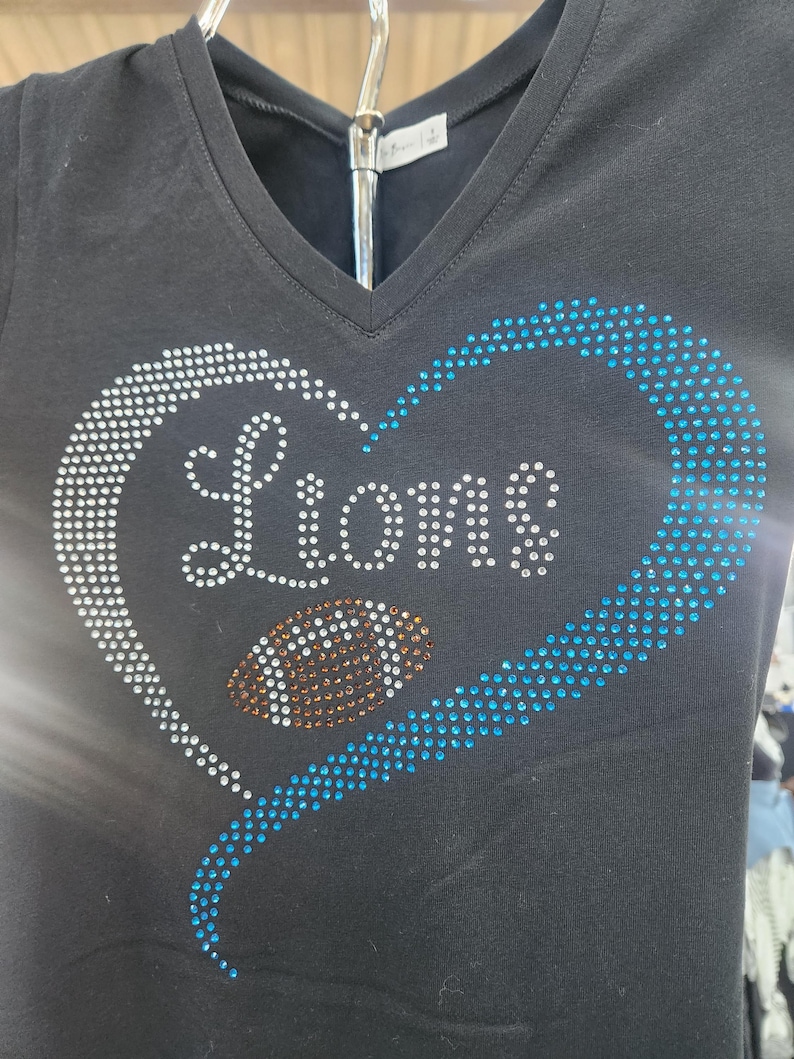 Może przedstawiać: Czarny t-shirt z dekoltem w serek z sercem z białych i niebieskich kryształk&oacute;w. Słowo "Lions" jest napisane białymi kryształkami, z kryształową piłką nożną pośrodku. Odzież o tematyce sportowej.