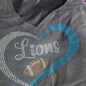 Może przedstawiać: Czarny t-shirt z dekoltem w serek z motywem w kształcie serca. Serce obrysowane jest białymi i niebieskimi kryształkami, z napisem "LIONS" z białych kryształk&oacute;w. W środku serca znajduje się kształt piłki nożnej z pomarańczowych kryształk&oacute;w.