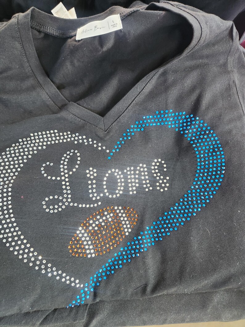 Może przedstawiać: Czarny t-shirt z dekoltem w serek z motywem w kształcie serca. Serce obrysowane jest białymi i niebieskimi kryształkami, z napisem "LIONS" z białych kryształk&oacute;w. W środku serca znajduje się kształt piłki nożnej z pomarańczowych kryształk&oacute;w.