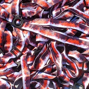 Könnte beinhalten: Ein Stapel rot-weißer Batik-Lanyards mit schwarzer Beschriftung. Die Lanyards haben eine schwarze Kunststoffschnalle.