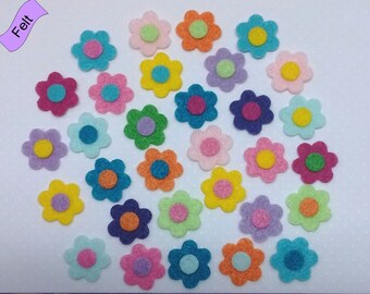 May include: A collection of small, multi-colored felt flower cutouts in various shades of pink, blue, yellow, orange, green, and purple, each featuring a contrasting center circle. A purple ruler is positioned at the bottom for scale.