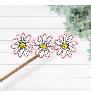 May include: Three daisy flower stickers with white petals, yellow centers, and pink outlines are attached to a wooden stick. The daisies are arranged in a row against a white wooden background.