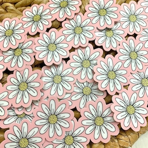 May include: A collection of daisy stickers with white petals, yellow centers, and a light pink background. The stickers are scattered on a woven, light brown surface. The daisy stickers are a popular craft item.