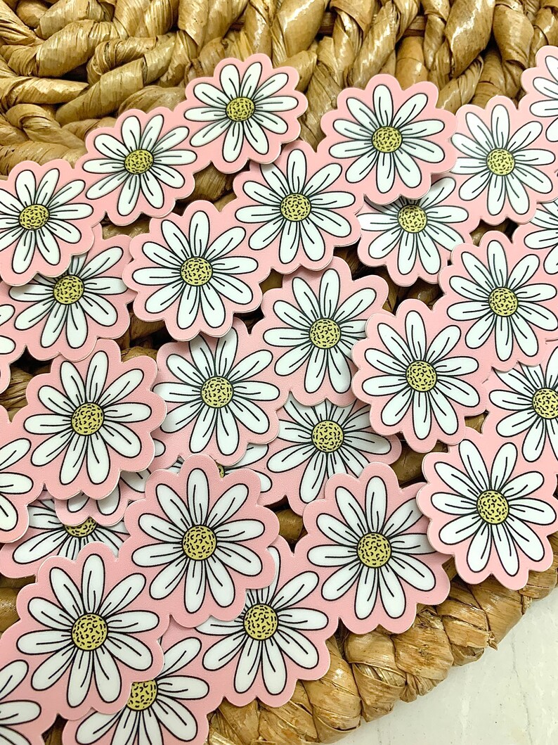 May include: A collection of daisy stickers with white petals, yellow centers, and a light pink background. The stickers are scattered on a woven, light brown surface. The daisy stickers are a popular craft item.