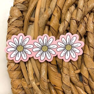 May include: Three daisy-shaped stickers with white petals, yellow centers, and a pink background. The stickers are arranged in a row and are set against a woven, light brown background.