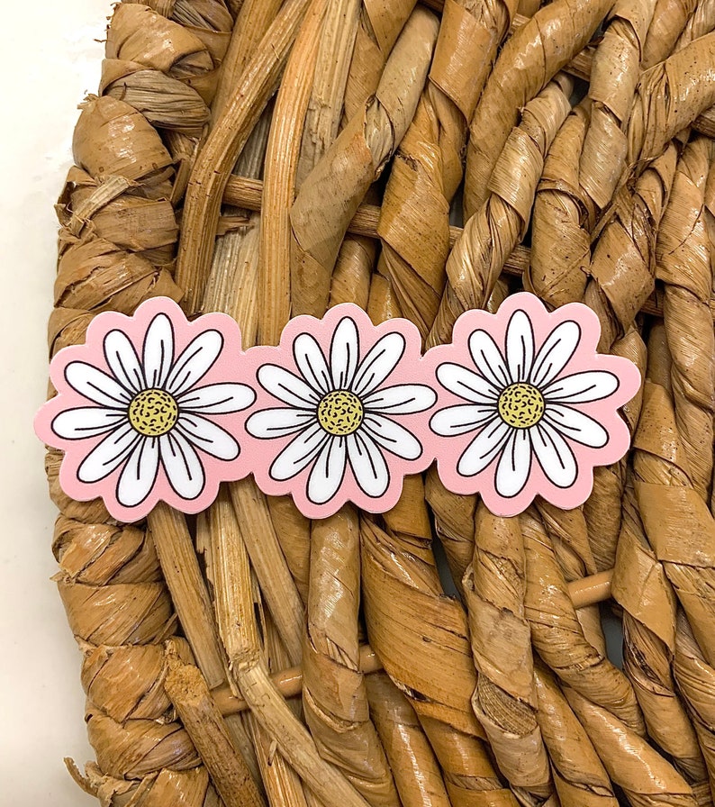 May include: Three daisy-shaped stickers with white petals, yellow centers, and a pink background. The stickers are arranged in a row and are set against a woven, light brown background.