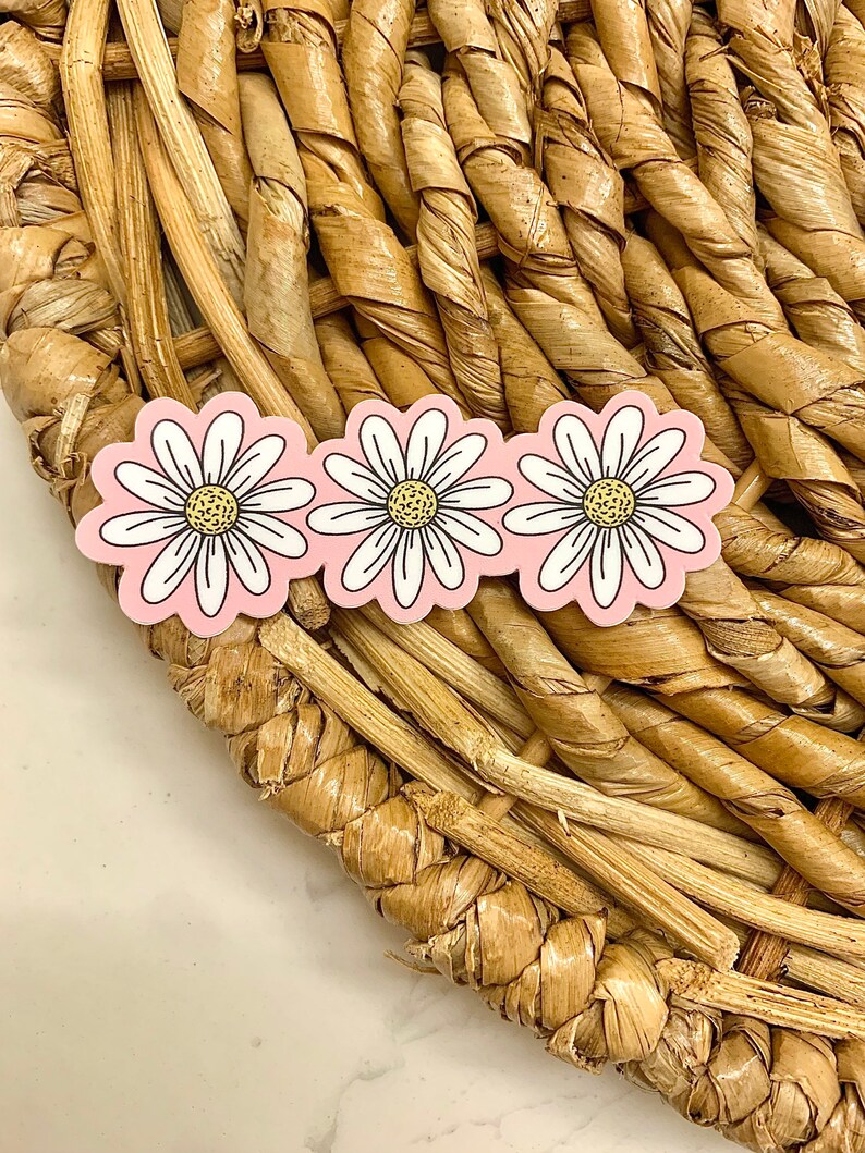May include: Three daisy-shaped stickers with white petals, yellow centers, and black outlines, set against a light pink background. The stickers are arranged in a row and are placed on a woven, light brown surface.