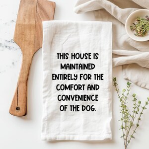 May include: White kitchen towel with the text "THIS HOUSE IS MAINTAINED ENTIRELY FOR THE COMFORT AND CONVENIENCE OF THE DOG." A wooden spatula and small bowl with herbs are also visible.