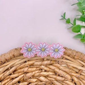 May include: Three daisy-shaped stickers with pink petals, white outlines, and yellow centers. The stickers are arranged horizontally on a woven, light brown surface. Green foliage is visible in the background.