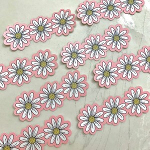 May include: Daisy flower stickers with white petals, yellow centers, and black outlines arranged in rows on a pink background. The stickers are die-cut and ready to peel and stick. Great for scrapbooking, crafts, or decorating.