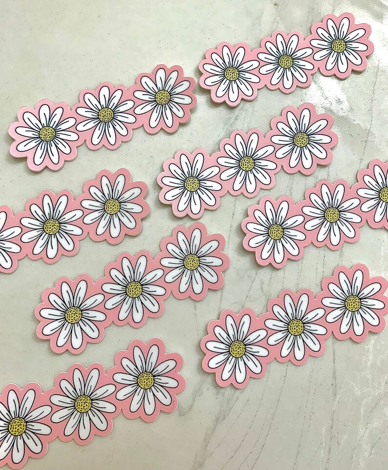 May include: Daisy flower stickers with white petals, yellow centers, and black outlines arranged in rows on a pink background. The stickers are die-cut and ready to peel and stick. Great for scrapbooking, crafts, or decorating.