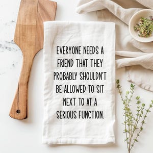 May include: White kitchen towel with the text "EVERYONE NEEDS A FRIEND THAT THEY PROBABLY SHOULDN'T BE ALLOWED TO SIT NEXT TO AT A SERIOUS FUNCTION." A wooden spatula and small bowl with herbs are also visible.