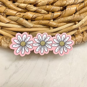 May include: A pink daisy chain with three white daisies with yellow centers and black outlines. The daisy chain is set against a woven background.