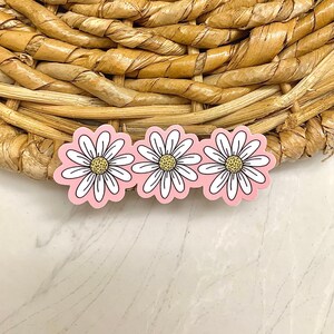 May include: Three daisy-shaped stickers with white petals, yellow centers, and black outlines are arranged in a row. The stickers have a pink background. The stickers are on a woven, light brown surface.