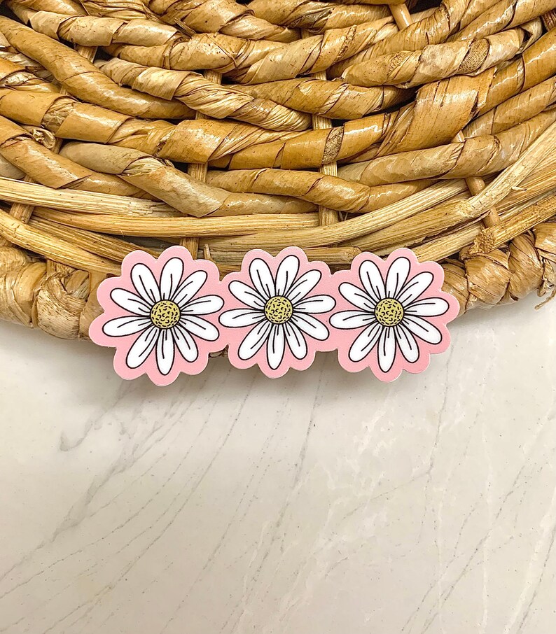 May include: Three daisy-shaped stickers with white petals, yellow centers, and black outlines are arranged in a row. The stickers have a pink background. The stickers are on a woven, light brown surface.