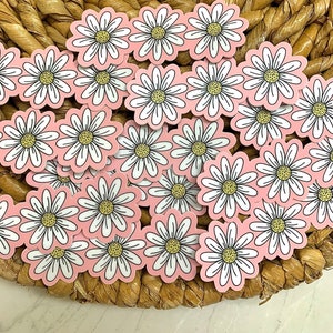 May include: A collection of daisy flower stickers with white petals, yellow centers, and black outlines, set against a light pink background. The stickers are arranged on a woven, brown surface.