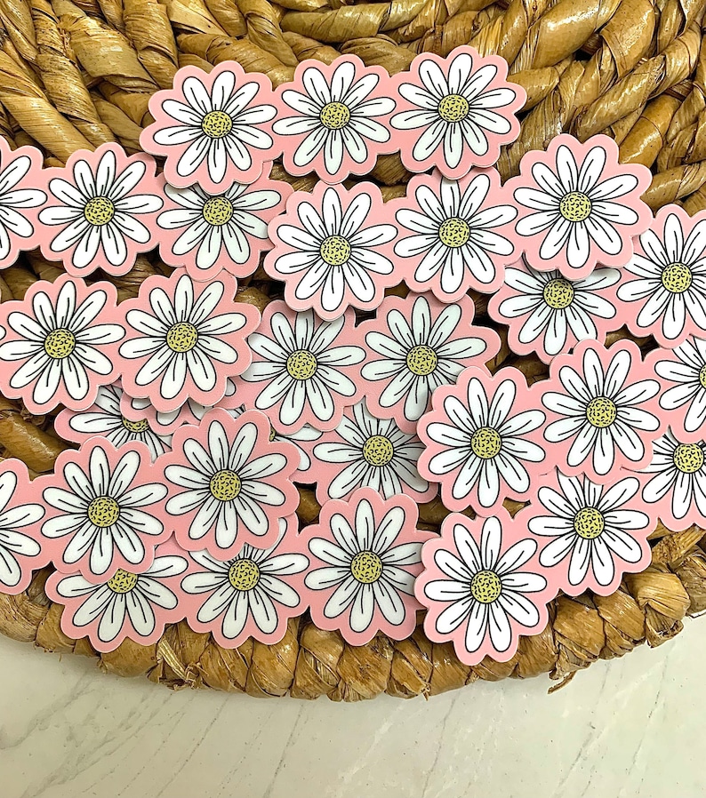 May include: A collection of daisy flower stickers with white petals, yellow centers, and black outlines, set against a light pink background. The stickers are arranged on a woven, brown surface.