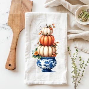 May include: A white kitchen towel with a hand-painted illustration of three pumpkins stacked in a blue and white porcelain vase. The pumpkins are orange, white, and cream with green stems and leaves.