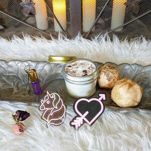Könnte beinhalten: Ein silbernes Tablett mit einer weißen Kerze in einem Glasgefäß, zwei braunen Kugeln, eine violette Flasche mit einem Einhorn-Anhänger, einem rosa und gelben Einhorn-Aufkleber und einem rosa Herz mit einem Pfeil-Aufkleber.