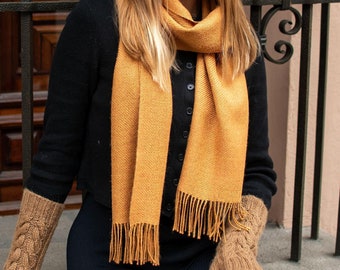 May include: A tan cable-knit beanie, a mustard yellow scarf with fringe ends, tan cable-knit fingerless gloves, and matching tan cable-knit leg warmers. The set is worn with a black button-down cardigan and black leggings.