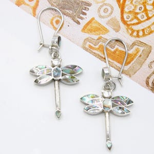 Peut inclure: Une paire de boucles d'oreilles en forme de libellule argentée avec des incrustations de nacre irisée sur les ailes et une petite pierre bleue au centre du corps. Les boucles d'oreilles pendent à des crochets simples et incluent une petite pierre.