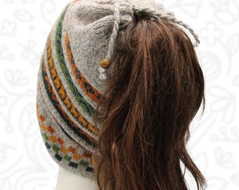 May include: A beige knit beanie with a drawstring opening at the crown, featuring a patterned band of orange, green, and dark brown geometric shapes. The hat includes a small wooden bead on the drawstring cord, designed to accommodate a ponytail.