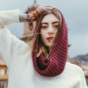 May include: A woman wearing a white knitted sweater with a burgundy infinity scarf and a colorful knitted fingerless glove.