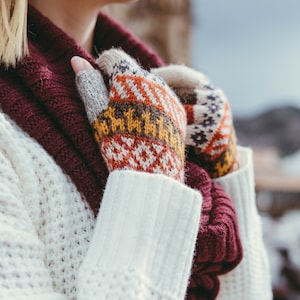 Puede incluir: Primer plano de una persona que lleva un suéter de punto blanco y una bufanda color burdeos. La persona lleva guantes sin dedos de punto de colores.