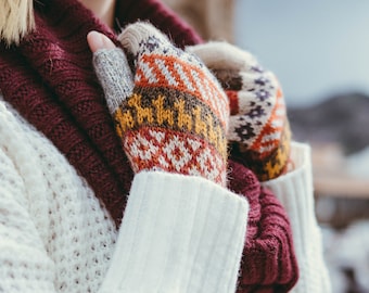 May include: Fingerless gloves featuring a multi-colored geometric pattern in shades of orange, yellow, brown, and cream. The gloves are worn with a chunky white knit sweater and a thick, ribbed, deep burgundy scarf draped around the neck.