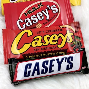 May include: Four Casey's candy bars with colorful packaging. The top bar is yellow with brown text, the second is red with white text, the third is red with yellow text, and the bottom bar is white with blue text. The bars are on a white, fluffy surface.