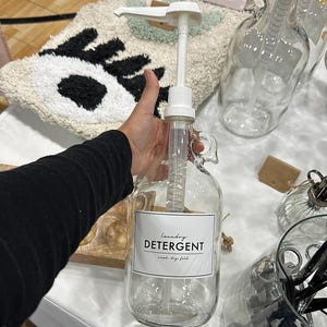 May include: A clear glass jug with a white plastic pump dispenser and a rectangular label that reads Laundry DETERGENT wash. dry. fold. The jug features a rounded body and a small handle near the top, designed for holding liquid soap or detergent.