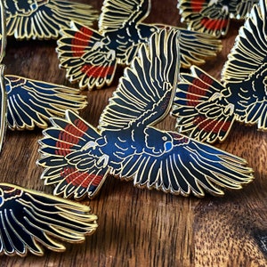 Op de afbeelding: Emaille pins met een zwart kaketoe-ontwerp met goudkleurige contouren. De vleugels van de vogel zijn uitgespreid en tonen rode en zwarte details. De pins zijn gerangschikt op een houten oppervlak.