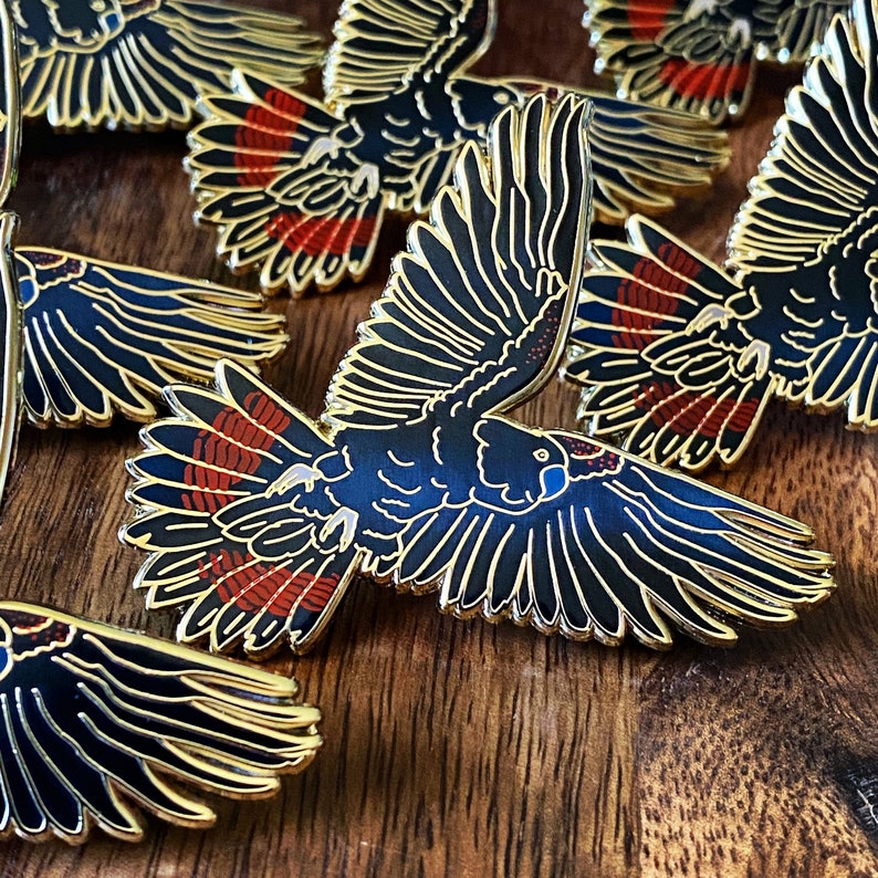 Op de afbeelding: Emaille pins met een zwart kaketoe-ontwerp met goudkleurige contouren. De vleugels van de vogel zijn uitgespreid en tonen rode en zwarte details. De pins zijn gerangschikt op een houten oppervlak.