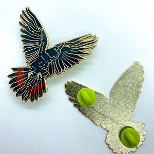 Op de afbeelding: Twee vogelvormige emaille pins. E&eacute;n pin toont de voorkant van de vogel met zwarte en gouden details en rode accenten op de staartveren. De andere pin toont de achterkant met een gouden afwerking en een groene pin back.