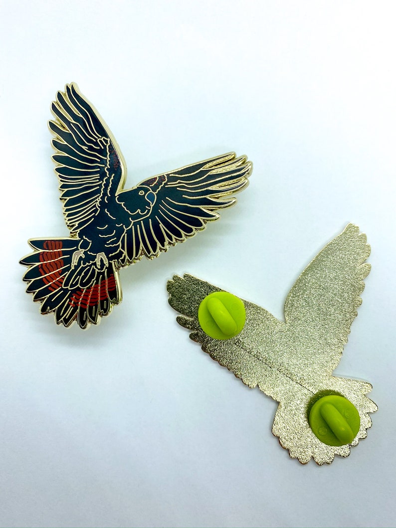 Op de afbeelding: Twee vogelvormige emaille pins. E&eacute;n pin toont de voorkant van de vogel met zwarte en gouden details en rode accenten op de staartveren. De andere pin toont de achterkant met een gouden afwerking en een groene pin back.