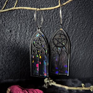 Könnte beinhalten: Schwarze Gothic-Ohrringe im Stained-Glass-Fenster-Design mit einem Pentagramm. Die Ohrringe haben schillernde Details und hängen an silberfarbenen Creolen. Der Hintergrund ist dunkel, und der Text "Black Rabbit White Rabbit" ist sichtbar.
