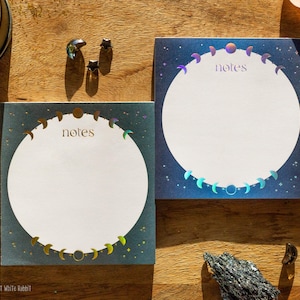 May include: Two square note pads featuring a central white circle surrounded by a holographic moon phase cycle and the word notes printed in the centre. The background is a dark blue gradient with small star accents, creating a celestial design.