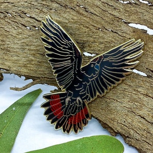 Op de afbeelding: Een zwarte en rode vogelspeld met goudkleurige randen, vleugels wijd gespreid. De speld is geplaatst tegen een getextureerde, bruine houten achtergrond met groene bladeren. De veren van de vogel zijn gedetailleerd met zwart en rood email.