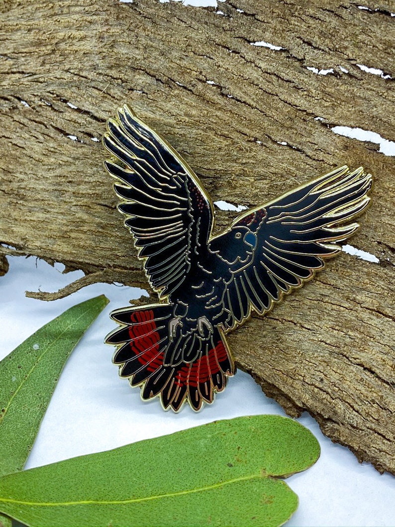 Op de afbeelding: Een zwarte en rode vogelspeld met goudkleurige randen, vleugels wijd gespreid. De speld is geplaatst tegen een getextureerde, bruine houten achtergrond met groene bladeren. De veren van de vogel zijn gedetailleerd met zwart en rood email.