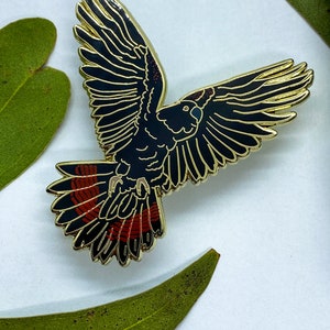 Op de afbeelding: Een zwarte en gouden emaille pin in de vorm van een vogel in vlucht. De vogel heeft rode accenten op zijn staartveren. De pin is geplaatst tegen een witte achtergrond met groene bladeren.