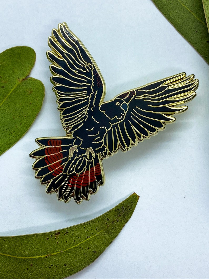 Op de afbeelding: Een zwarte en gouden emaille pin in de vorm van een vogel in vlucht. De vogel heeft rode accenten op zijn staartveren. De pin is geplaatst tegen een witte achtergrond met groene bladeren.