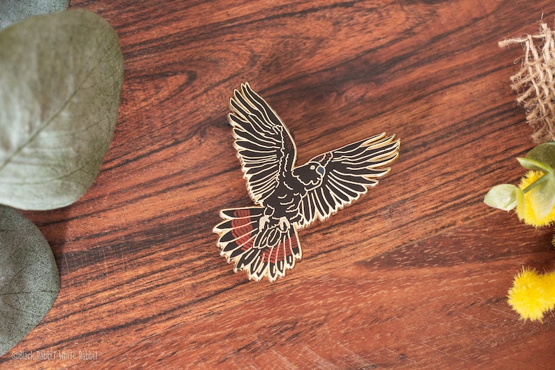 Op de afbeelding: Een goudkleurige vogelspeld met zwarte en rode details. De speld toont een vogel met uitgespreide vleugels, tegen een donkere houtnerf achtergrond. De veren van de vogel zijn gedetailleerd.