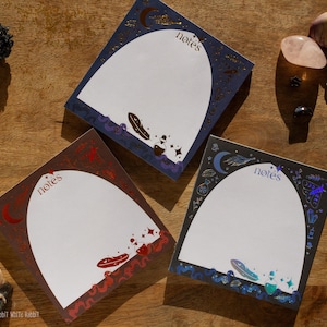 May include: Three square note pads with arched white writing areas, each featuring the word notes at the top. The borders are decorated with celestial and mystical illustrations including moons, feathers, and bottles in blue, red, and black tones.