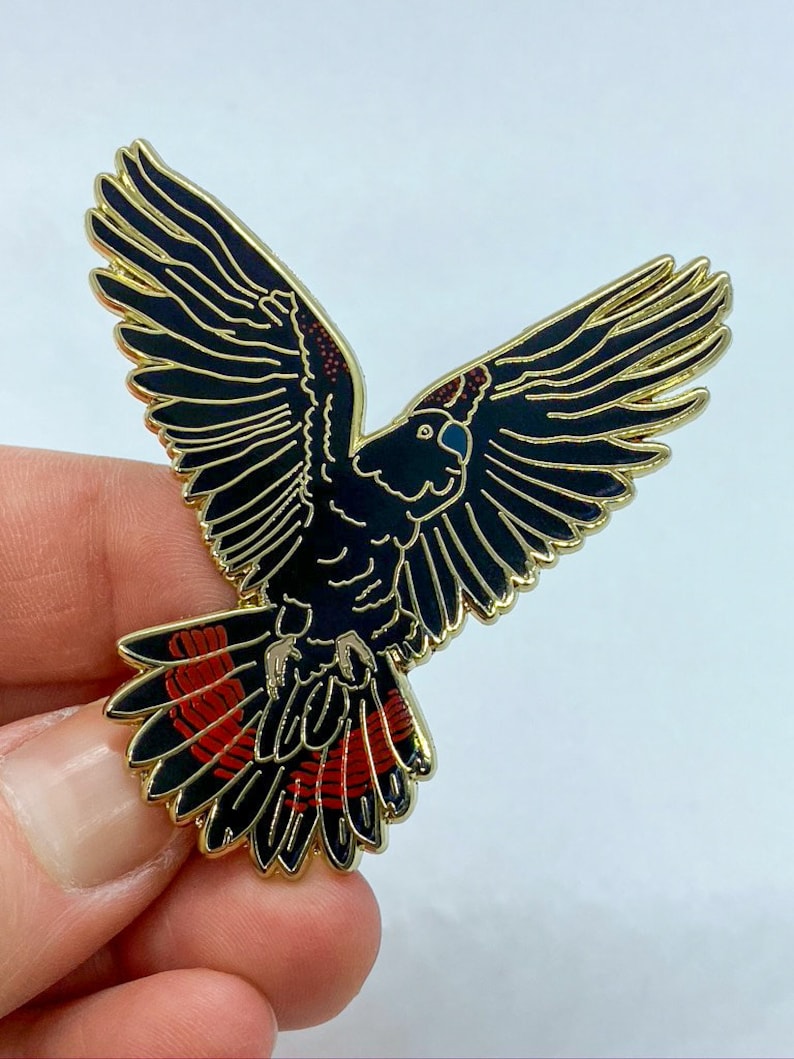 Op de afbeelding: Een zwarte en gouden emaille pin in de vorm van een vogel met uitgespreide vleugels. De vogel heeft rode accenten op zijn staartveren en kop. De pin heeft een gouden rand en staat tegen een witte achtergrond.